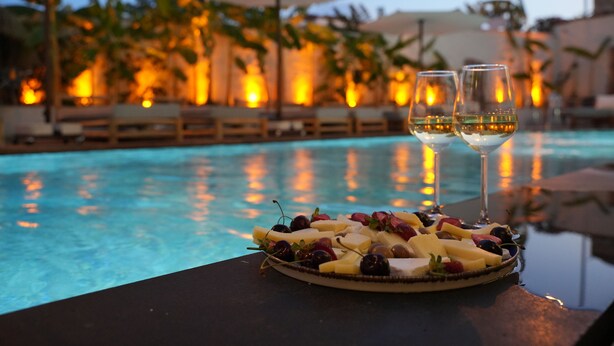 A poolside table with a fruit platter and two glasses of white wine, overlooking a pool with lounge chairs and umbrellas in the background. - Q Alacati Otel (Cesme)