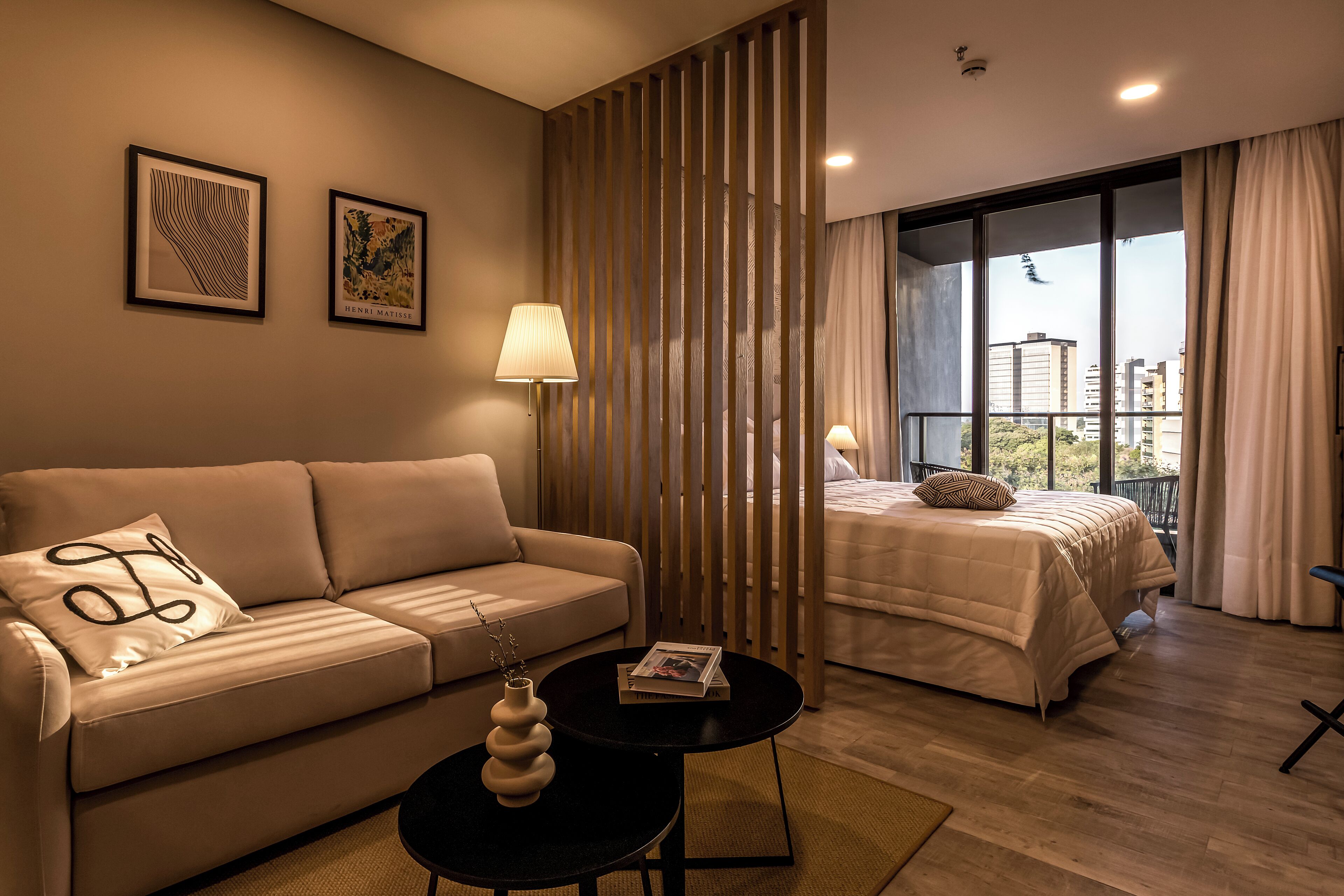 A modern hotel room with a sofa, a small table with a vase, a bed with a view of the city, and a decorative partition.