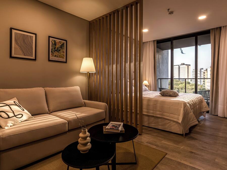 A modern hotel room with a sofa, a small table with a vase, a bed with a view of the city, and a decorative partition.