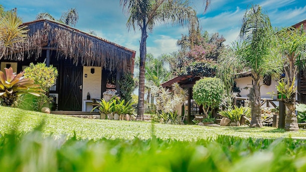 A tropical garden with a thatched-roof building, surrounded by lush greenery and palm trees. - Pousada Rosa da Praia - Praia do Rosa (Imbituba)