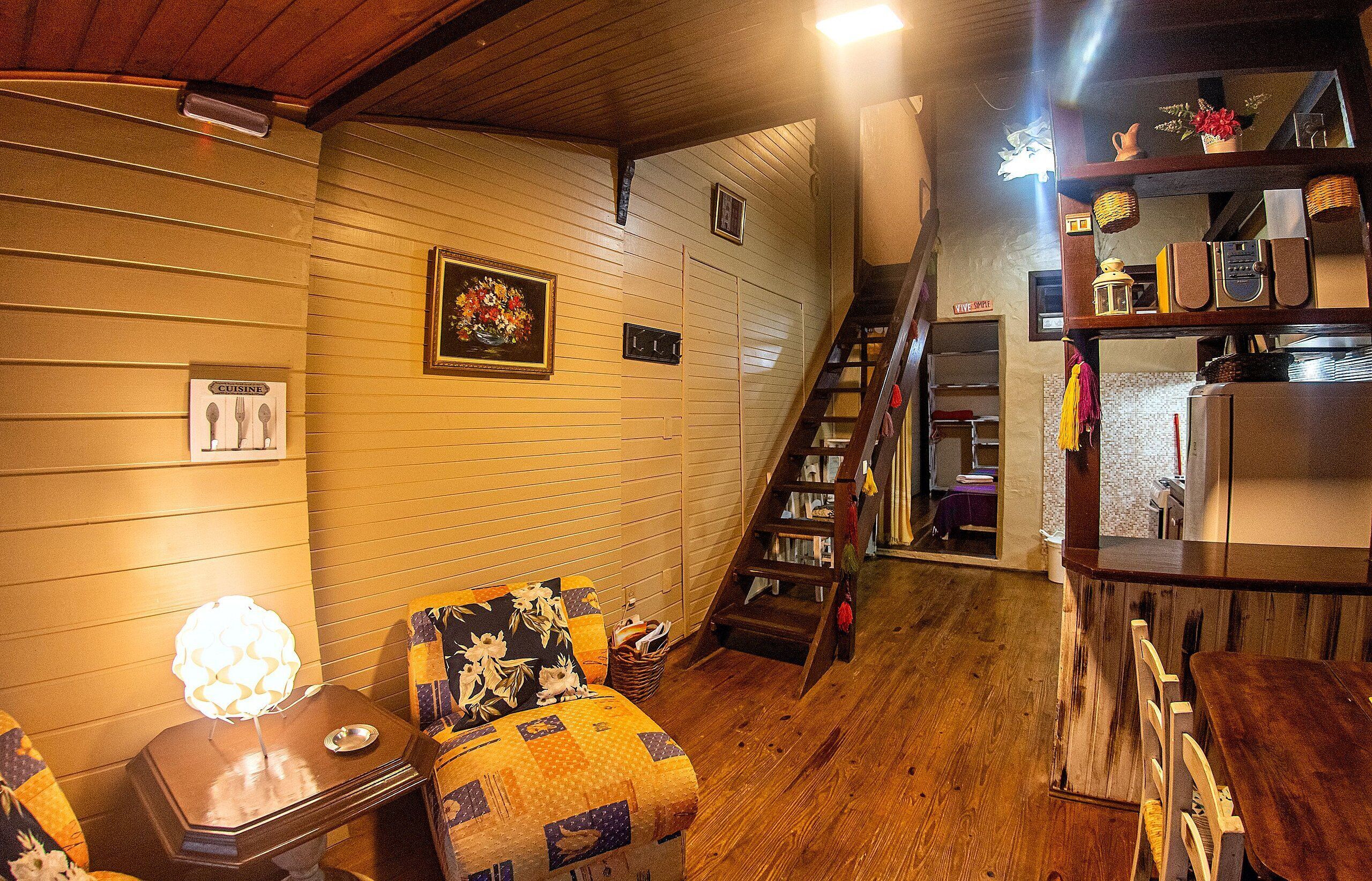 A cozy living room with a wooden floor, a quilted chair, a table with a lamp, and a staircase leading to another room.
