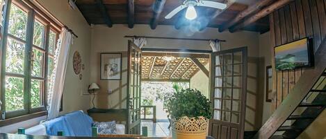 Ein Zimmer mit Holzboden, Deckenventilator, großem Fenster mit Vorhängen, Topfpflanze, Treppe und Fernseher an der Wand.