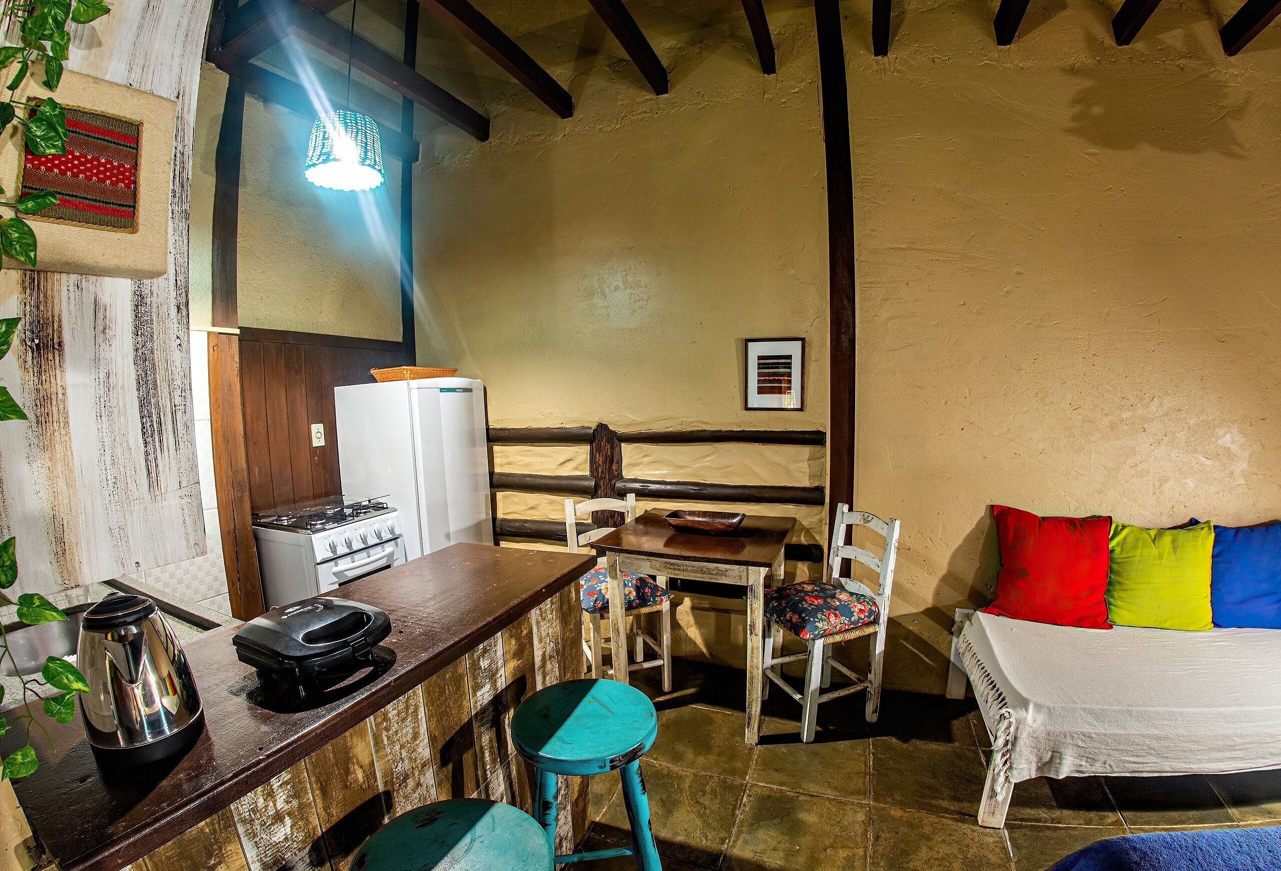 A cozy room with a wooden bar, a bed with colorful pillows, a small table with chairs, a kettle, and a stove.