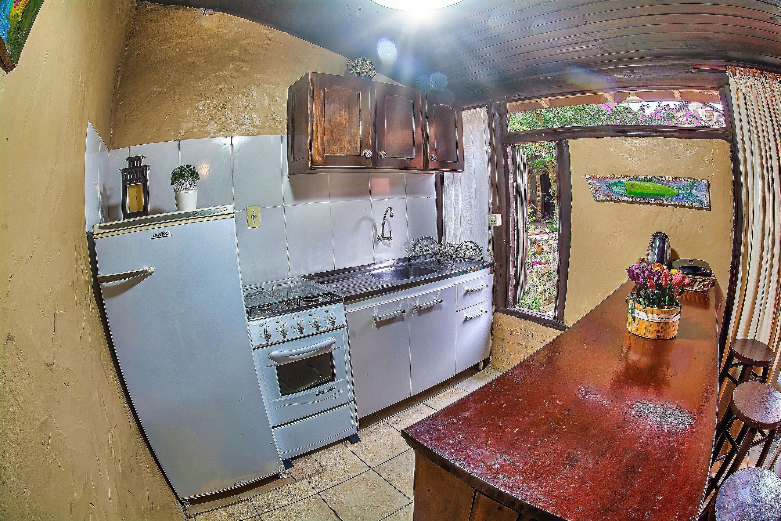 Cocina pequeña con armarios blancos, estufa y lavamanos. Hay una mesa de madera con sillas y una ventana con cortinas.