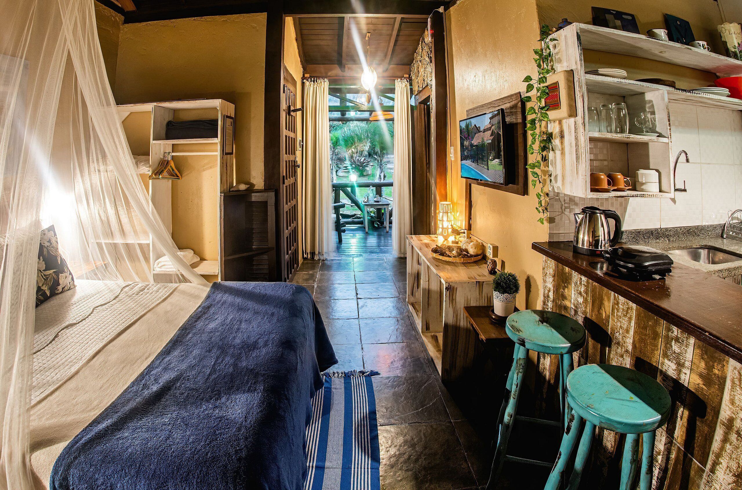Un pasillo estrecho que conduce a un dormitorio con una cama, una mesita de noche con lámpara y una pequeña cocina equipada con taburetes y un fregadero.