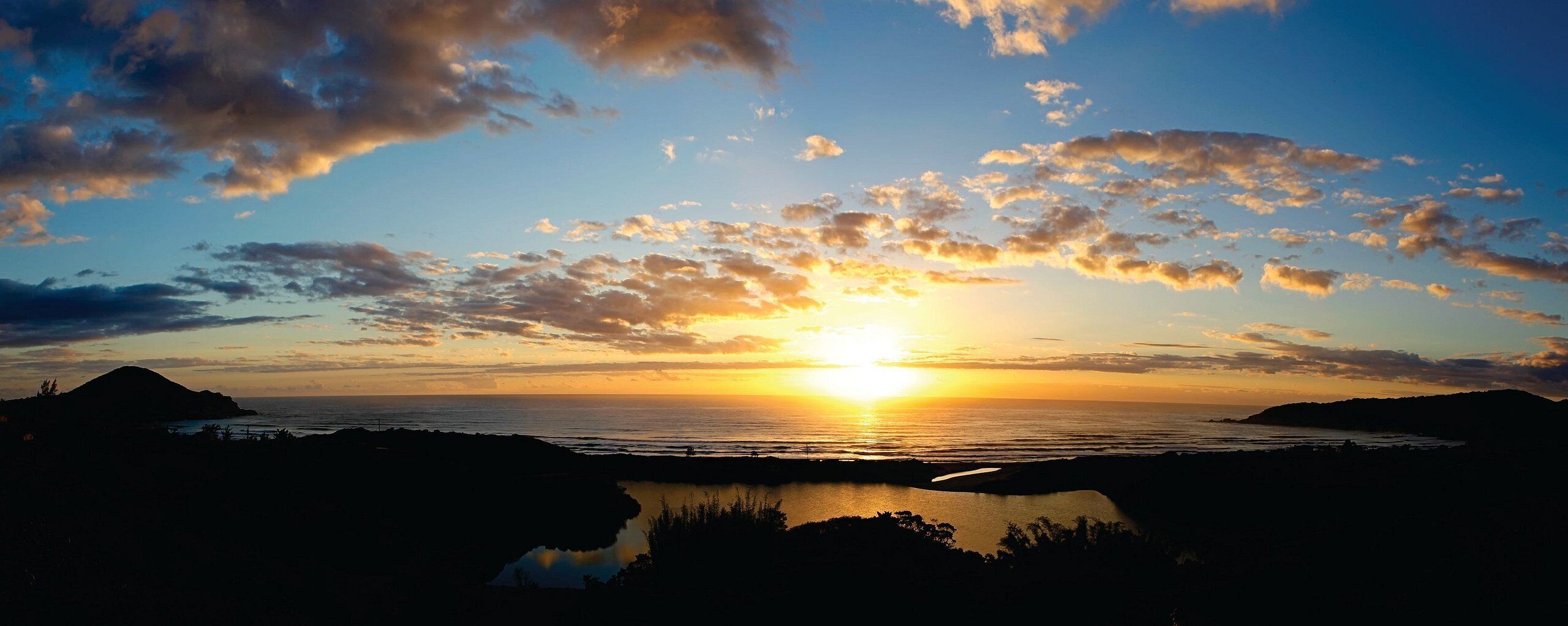 Um pôr do sol sobre uma paisagem costeira, com um corpo de água em primeiro plano e colinas ao fundo.