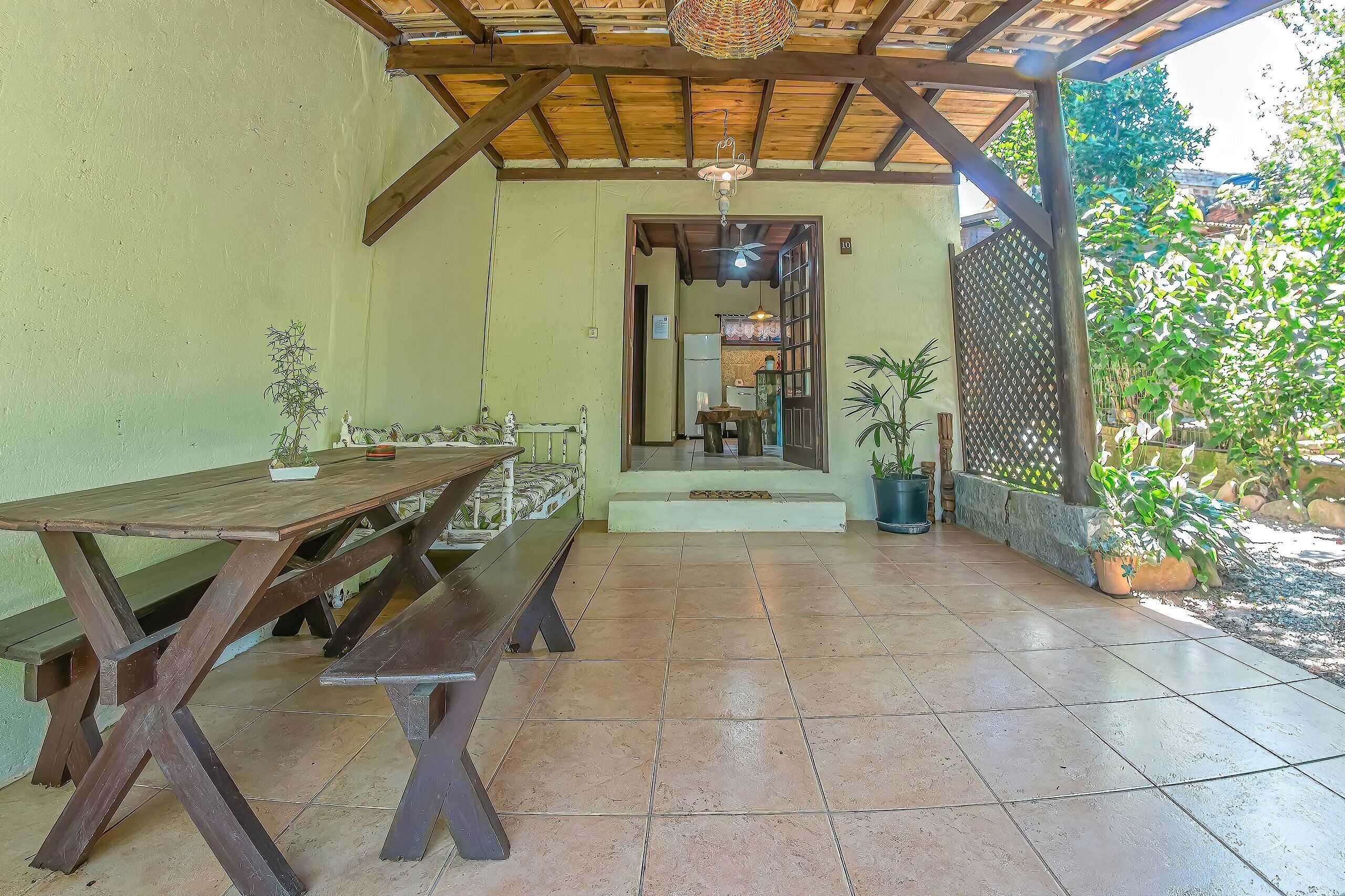 Uma varanda coberta com mesa e bancos de madeira, piso de azulejo e vasos com plantas.