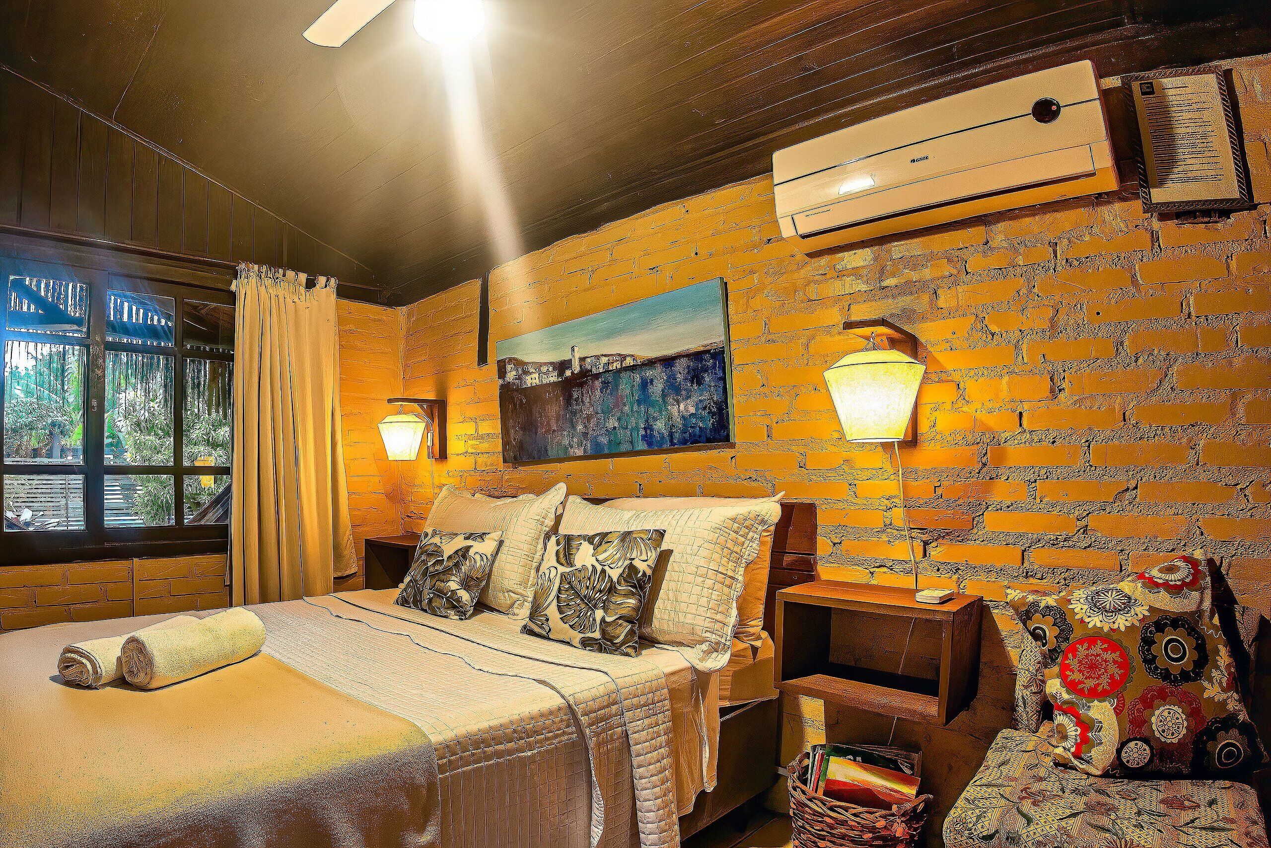 Una habitación de hotel con una pared de ladrillo, una cama con manta y almohadas, una mesita de noche, una silla y un cuadro en la pared.