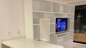Una cocina moderna con una encimera blanca, dos taburetes de bar, un televisor montado en una pared blanca y un armario de vidrio.
