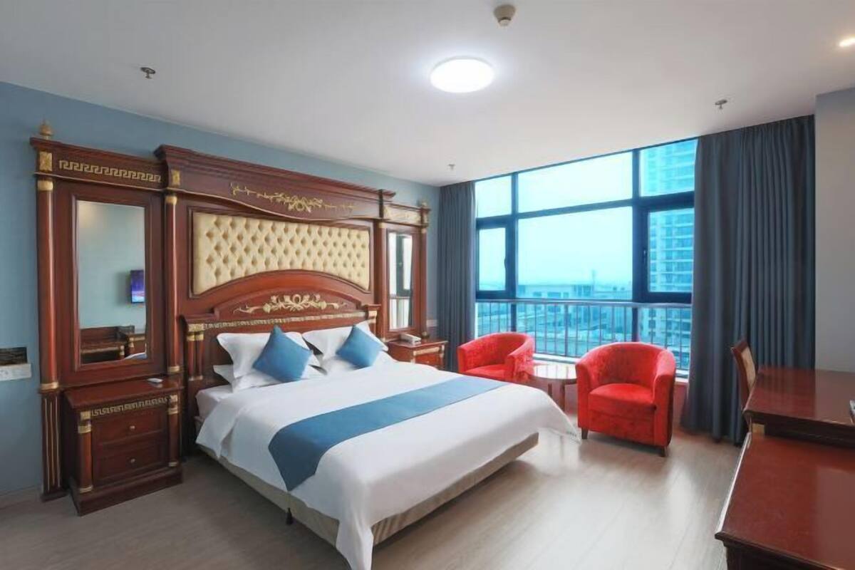 Une chambre d’hôtel avec un grand lit, une tête de lit en bois, un fauteuil rouge, une petite table et une grande fenêtre donnant sur la ville.