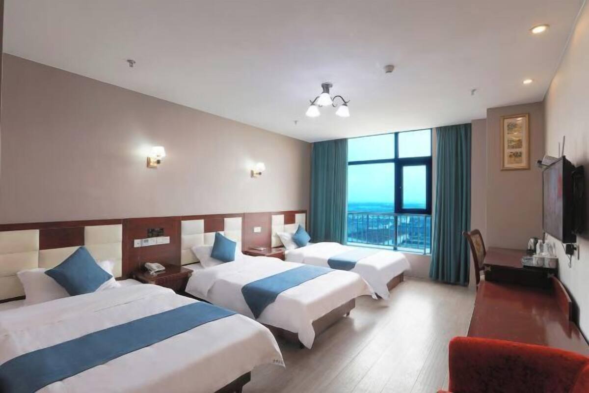 Une chambre d’hôtel avec trois lits, un bureau, une chaise, une télévision et une grande fenêtre donnant sur un beau paysage.