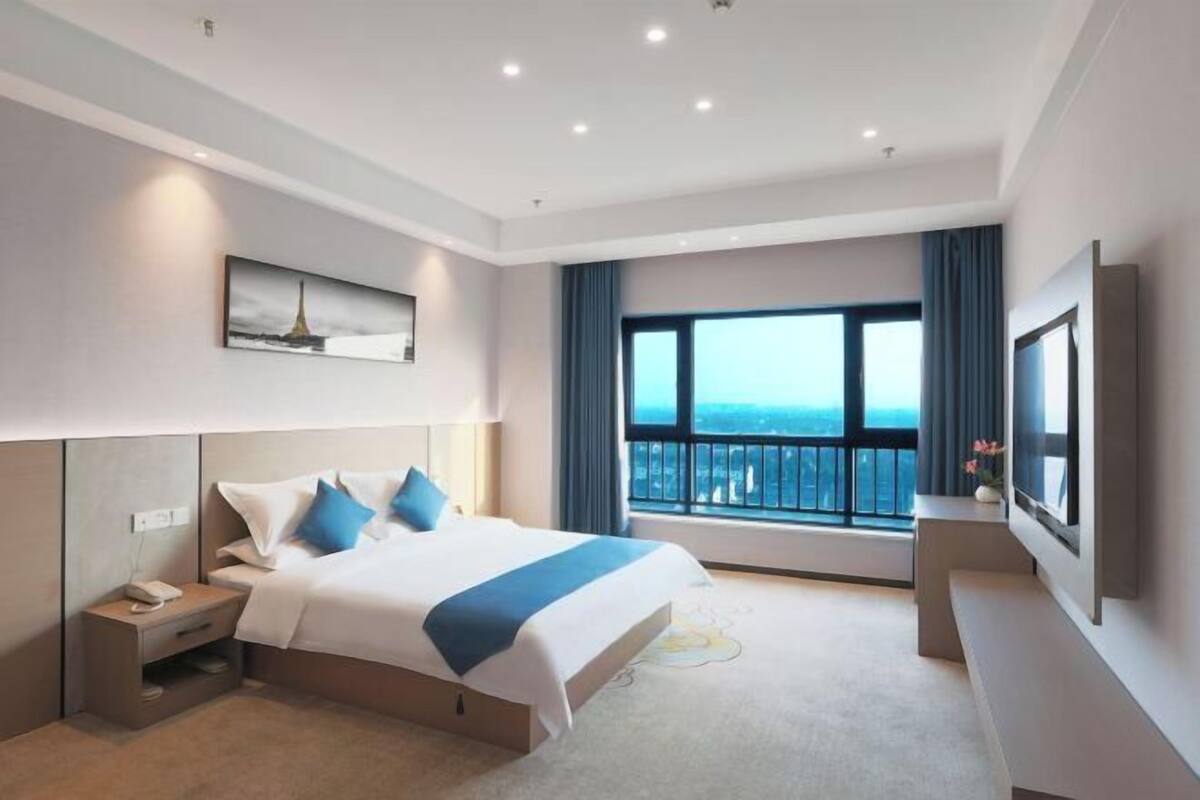 Une chambre d’hôtel moderne dotée d’un grand lit, d’une table de chevet avec un téléphone, d’une télévision à écran plat et d’une grande fenêtre offrant une vue sur la ville.