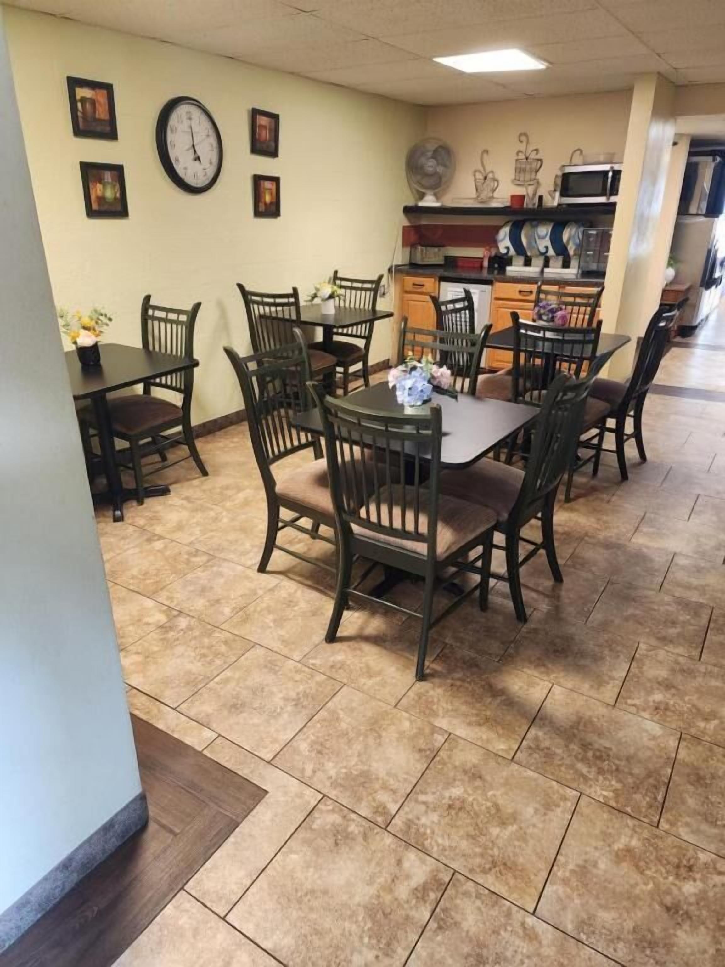 A dining area with multiple tables and chairs, a clock on the wall, and a kitchen area with appliances.