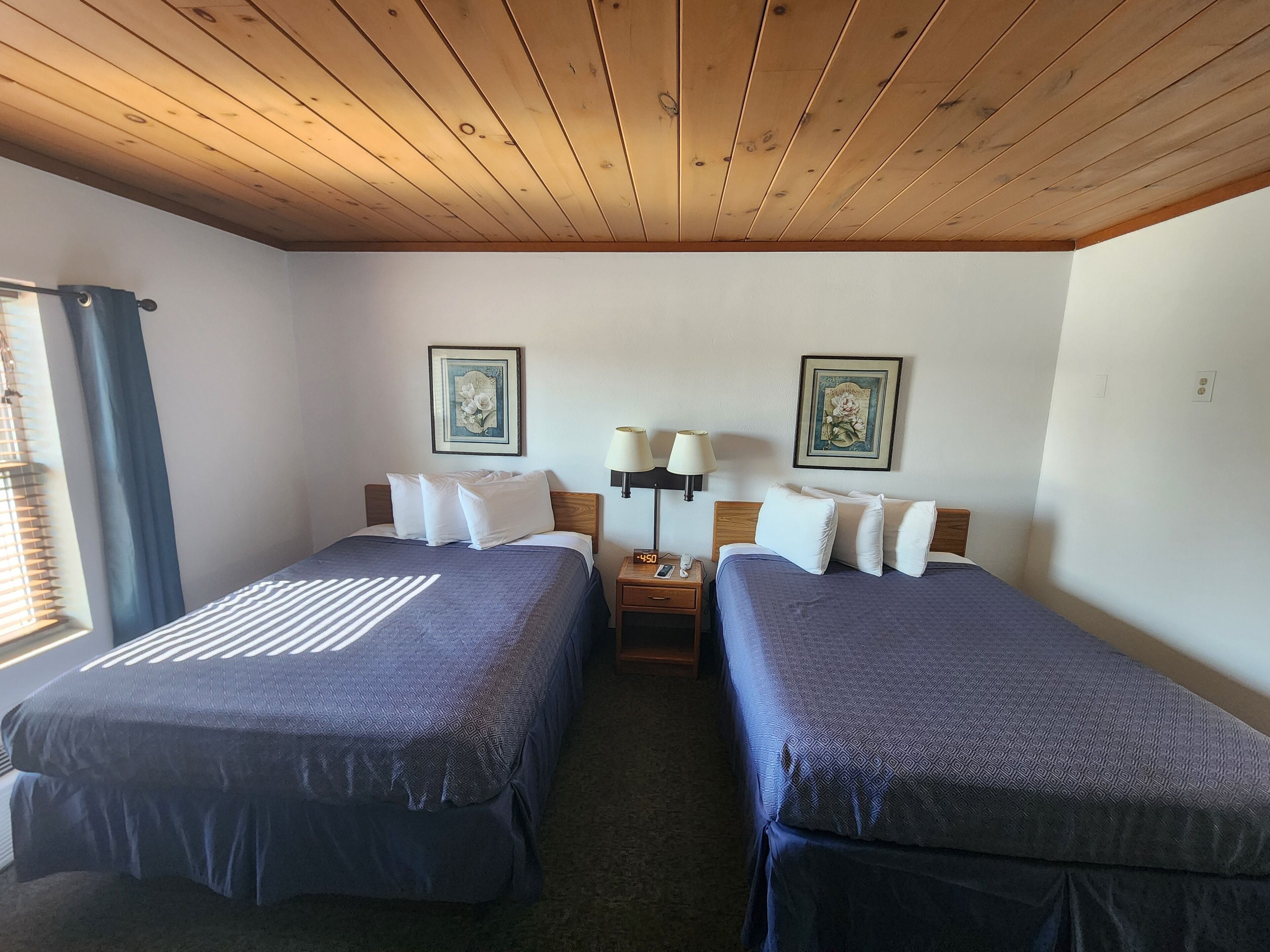 Habitación de hotel con dos camas, techo de madera, dos lámparas fijadas a la pared y dos cuadros enmarcados.