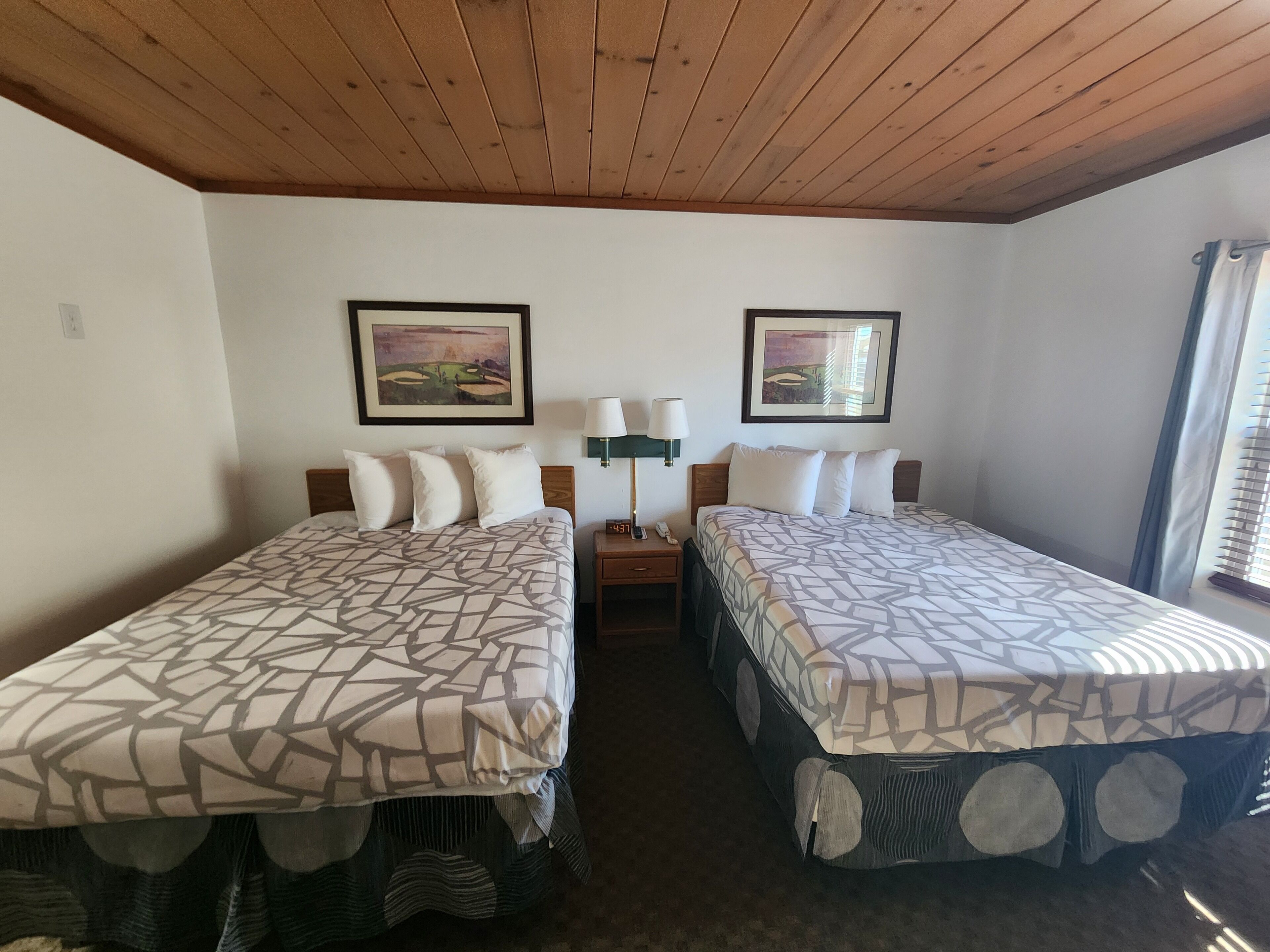 A hotel room with two beds, each with patterned bedding, a nightstand with a lamp, and framed artwork on the walls.