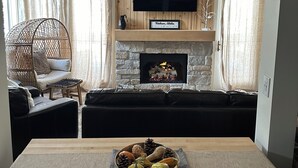 A living room with a stone fireplace, a flat-screen TV mounted above it, a dining table with a fruit bowl, and a black leather sofa. - Beautifully Remodeled Condo in Unbeatable Location! Walk to Gondola & Downtown (Ketchum)