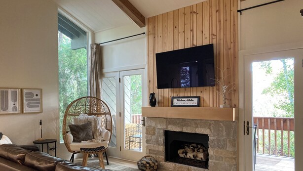 A living room with a stone fireplace, a large flat-screen TV mounted above it, a wicker chair, a leather sofa, and a view of the outdoors through a sliding glass door. - Beautifully Remodeled Condo in Unbeatable Location! Walk to Gondola & Downtown (Ketchum)