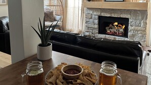 A cozy living room with a stone fireplace, a flat-screen TV mounted above it, and a comfortable seating area with a leather couch and a wicker chair. - Beautifully Remodeled Condo in Unbeatable Location! Walk to Gondola & Downtown (Ketchum)