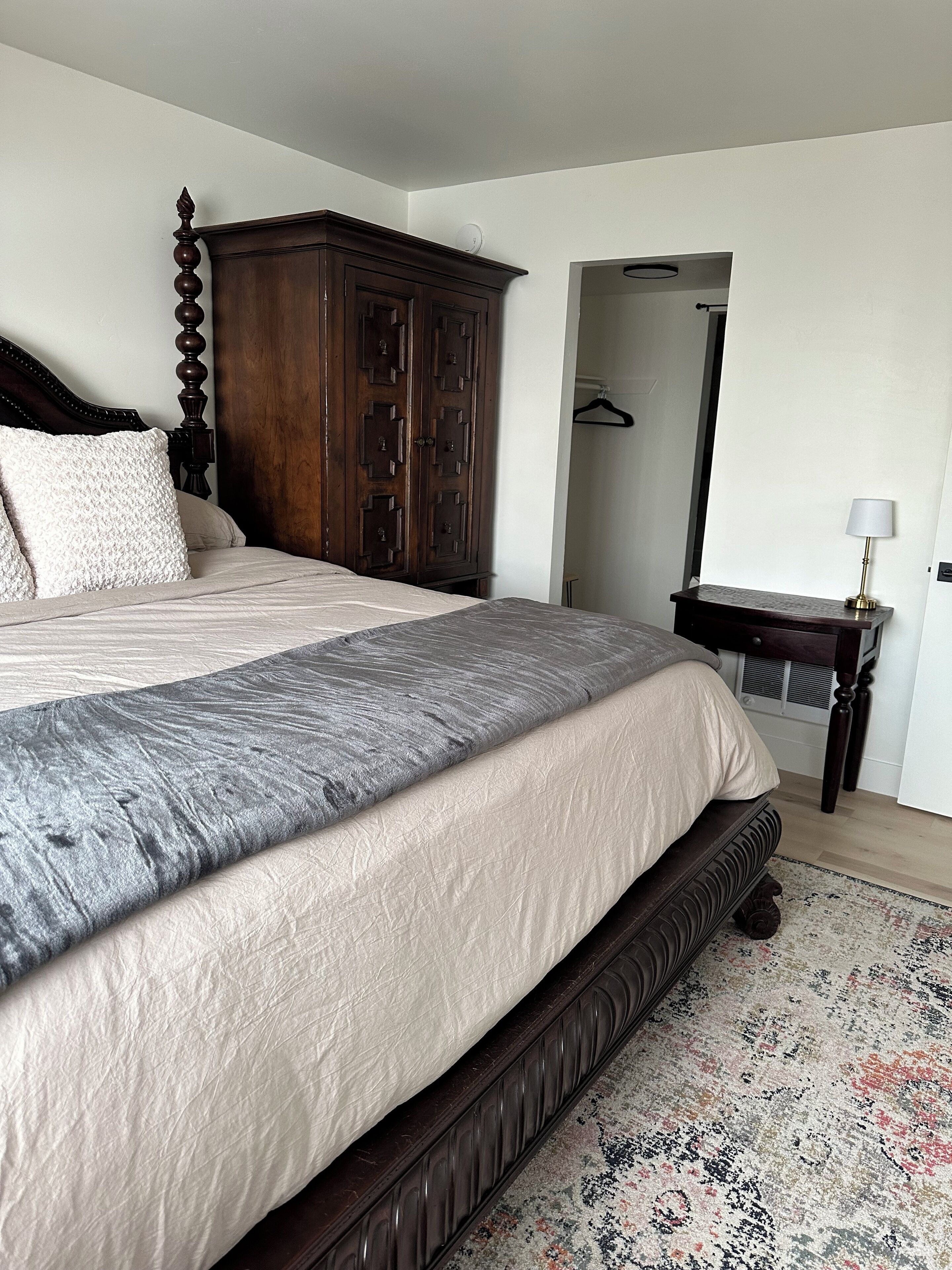 A bedroom with a large bed, a wooden wardrobe, a small table with a lamp, and a patterned rug.