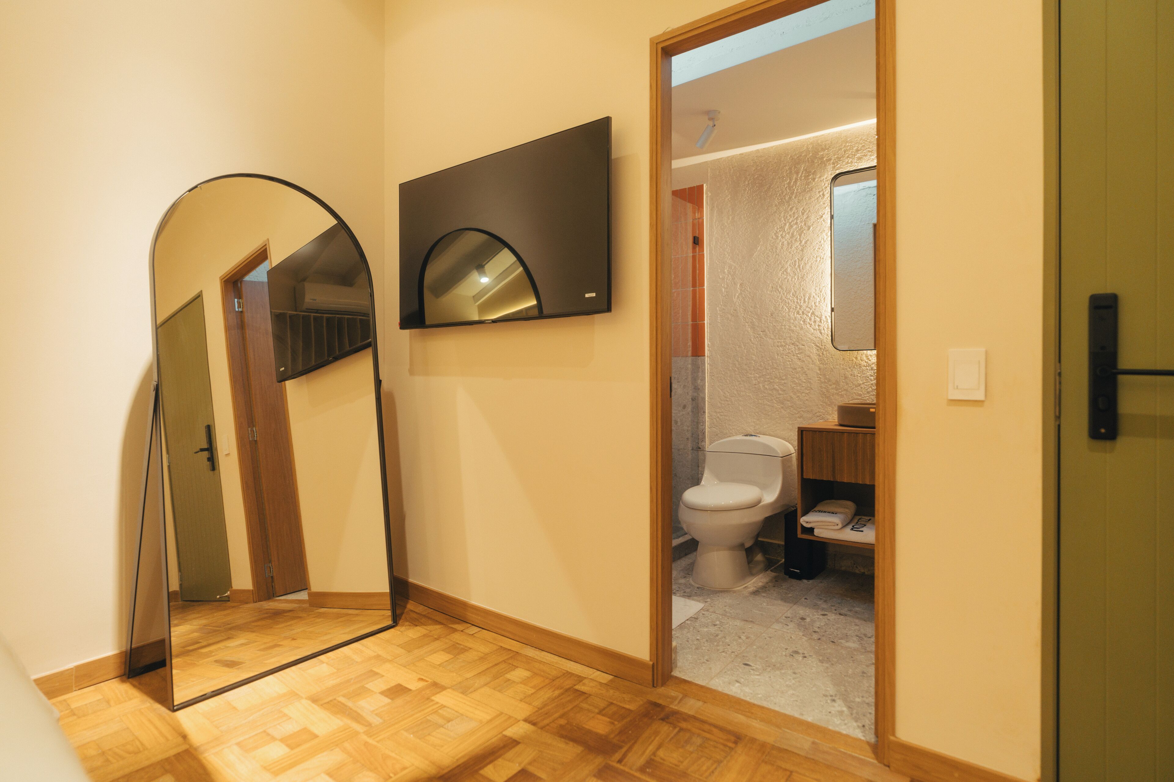 Een gang met een houten vloer, een spiegel en uitzicht op een badkamer met toilet en wastafel.