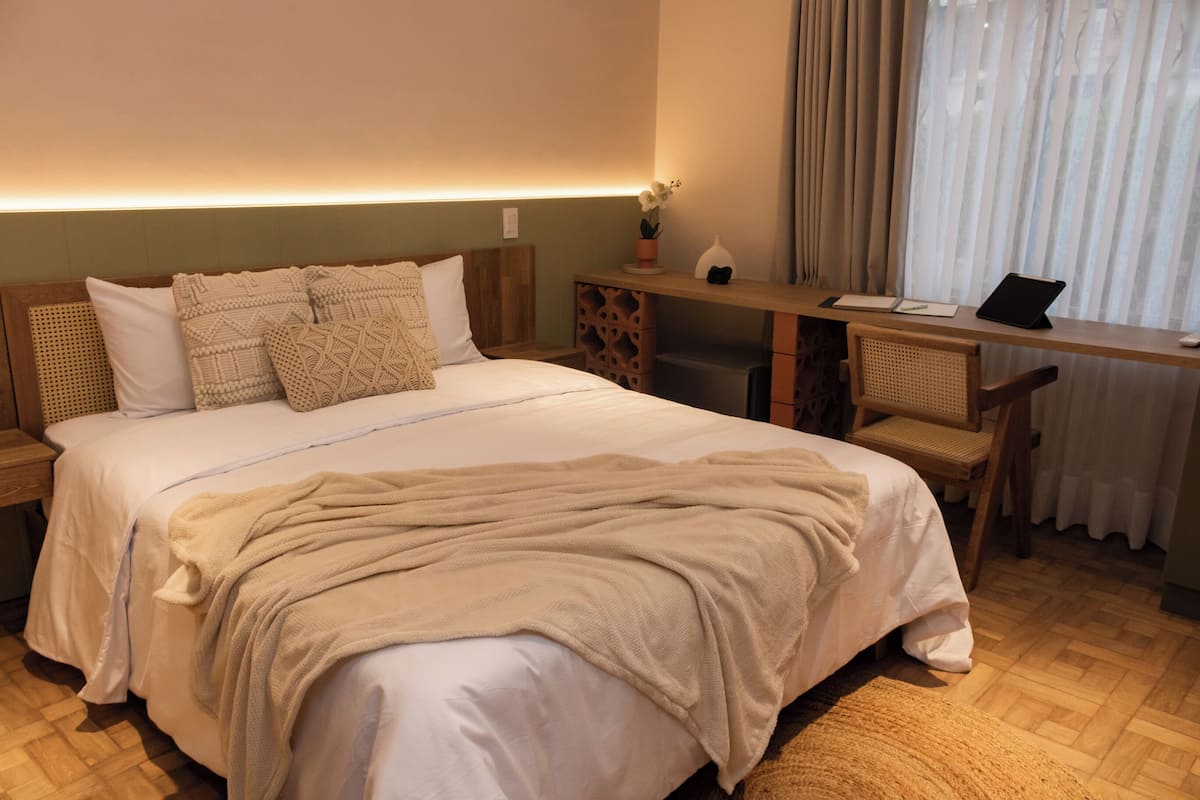 A neatly made bed with a beige blanket, a wooden headboard, and a bedside table with a lamp and a small potted plant.