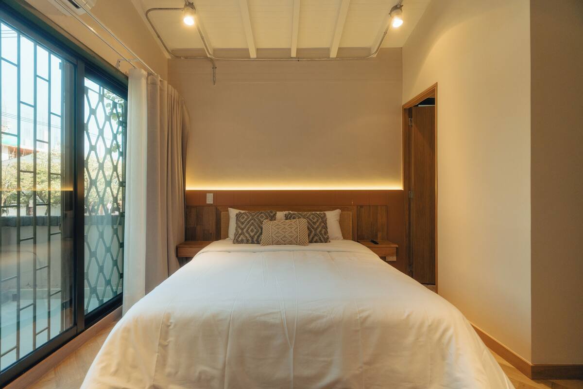 A bedroom with a large bed, a wooden headboard, a nightstand, and a window with a view of the outside.