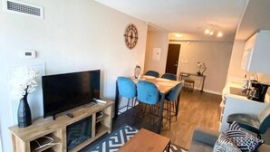A modern living room with a flat-screen TV, a wooden entertainment unit, a dining area with blue chairs, and a patterned rug. - GLOBALSTAY. Charming Lake View 1bd+den in Downtown (Toronto/Canada)