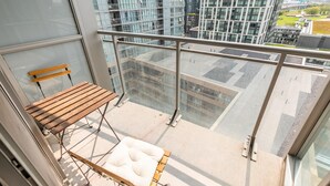 Een balkon met een houten bankje en een gewatteerd zitje, met uitzicht op een stadsgezicht met moderne gebouwen.