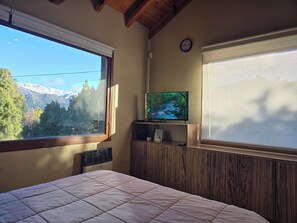 Une chambre à coucher avec un lit, un téléviseur posé sur un meuble en bois, une horloge au mur et une fenêtre donnant sur un paysage de montagnes et d’arbres.