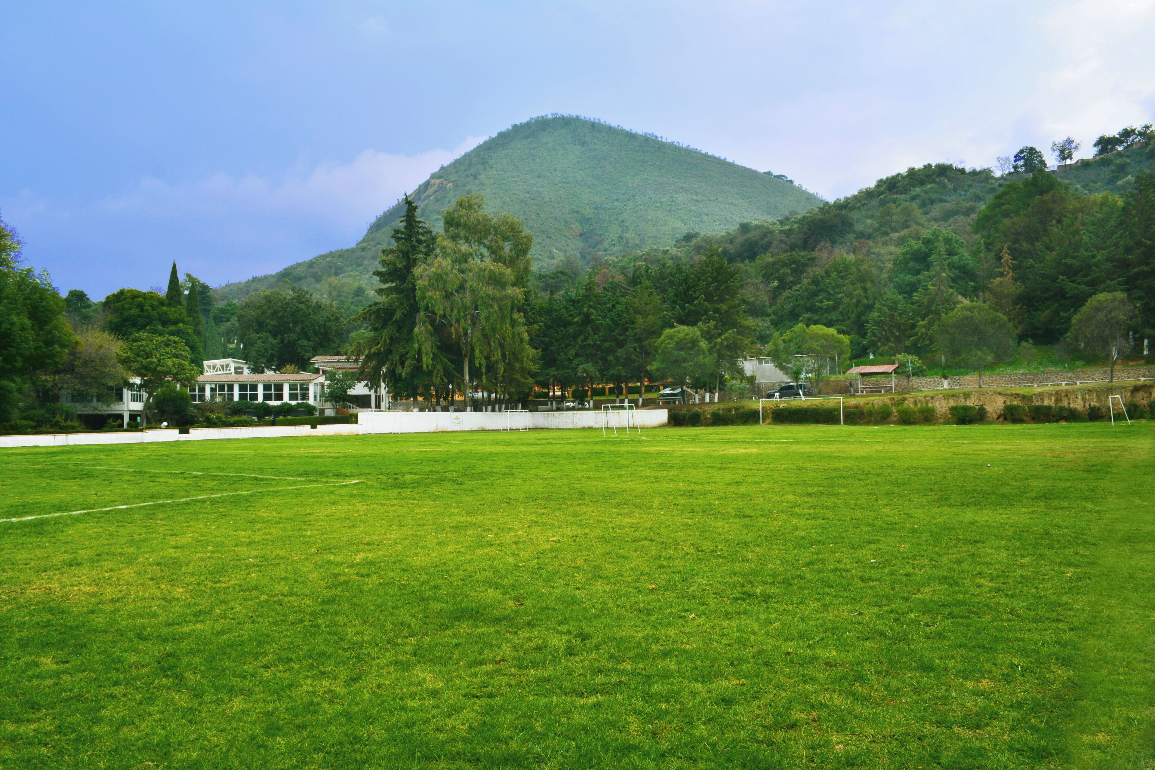 A grassy field with a mountain in the background, a building with a white facade, and a red-roofed structure.