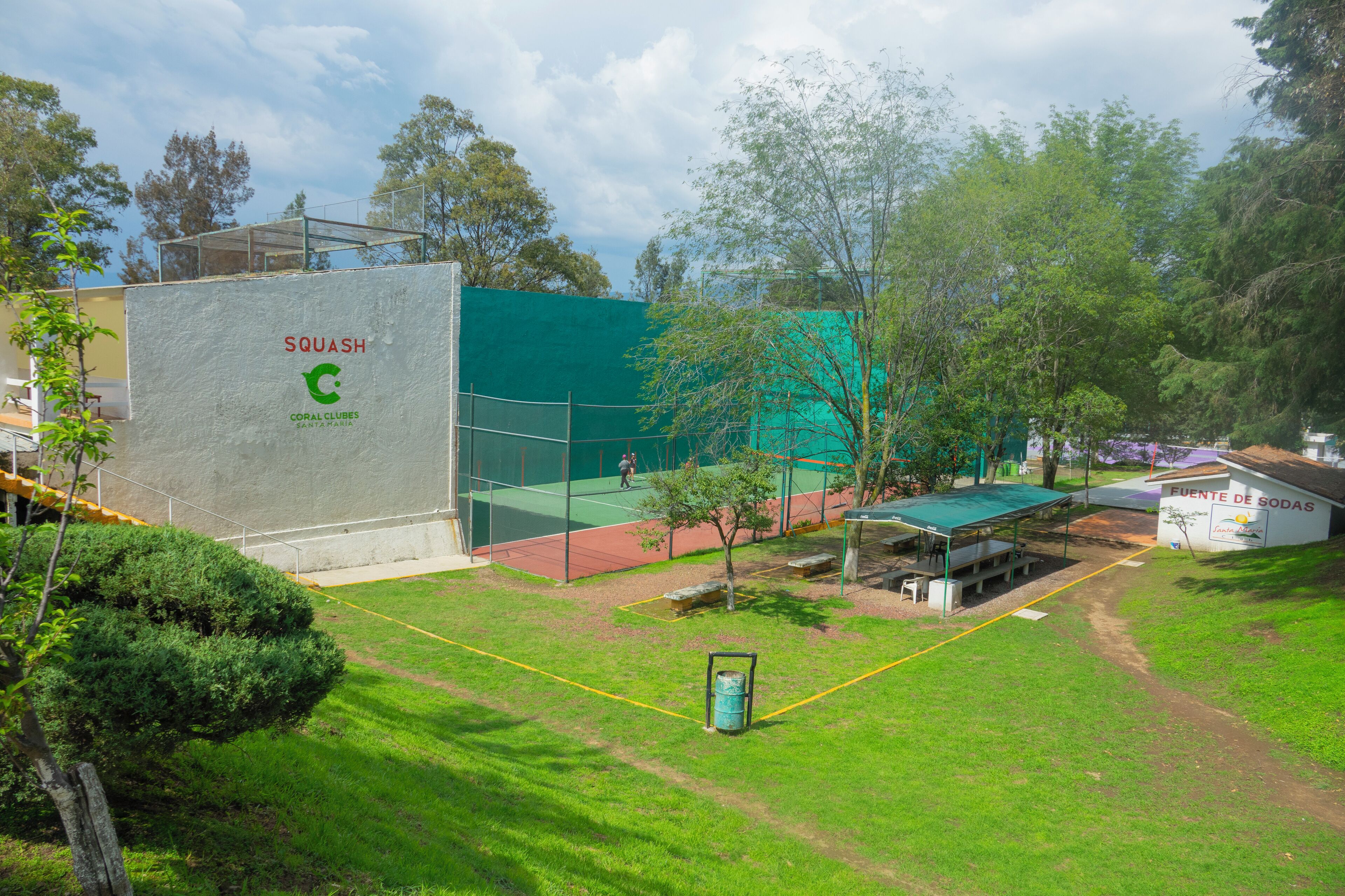 Un court de tennis entouré d’une clôture verte, avec un espace de repos couvert et un bâtiment portant l’inscription « SQUASH CORAL CLUBES SANTA MANIA ».