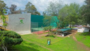 Un court de tennis entouré d’une clôture verte, avec un espace de repos couvert et un bâtiment portant l’inscription « SQUASH CORAL CLUBES SANTA MANIA ».