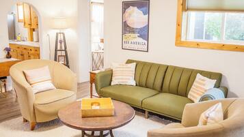 Sala de estar com sofá verde, poltronas bege, mesa de centro de madeira e um pôster emoldurado na parede.