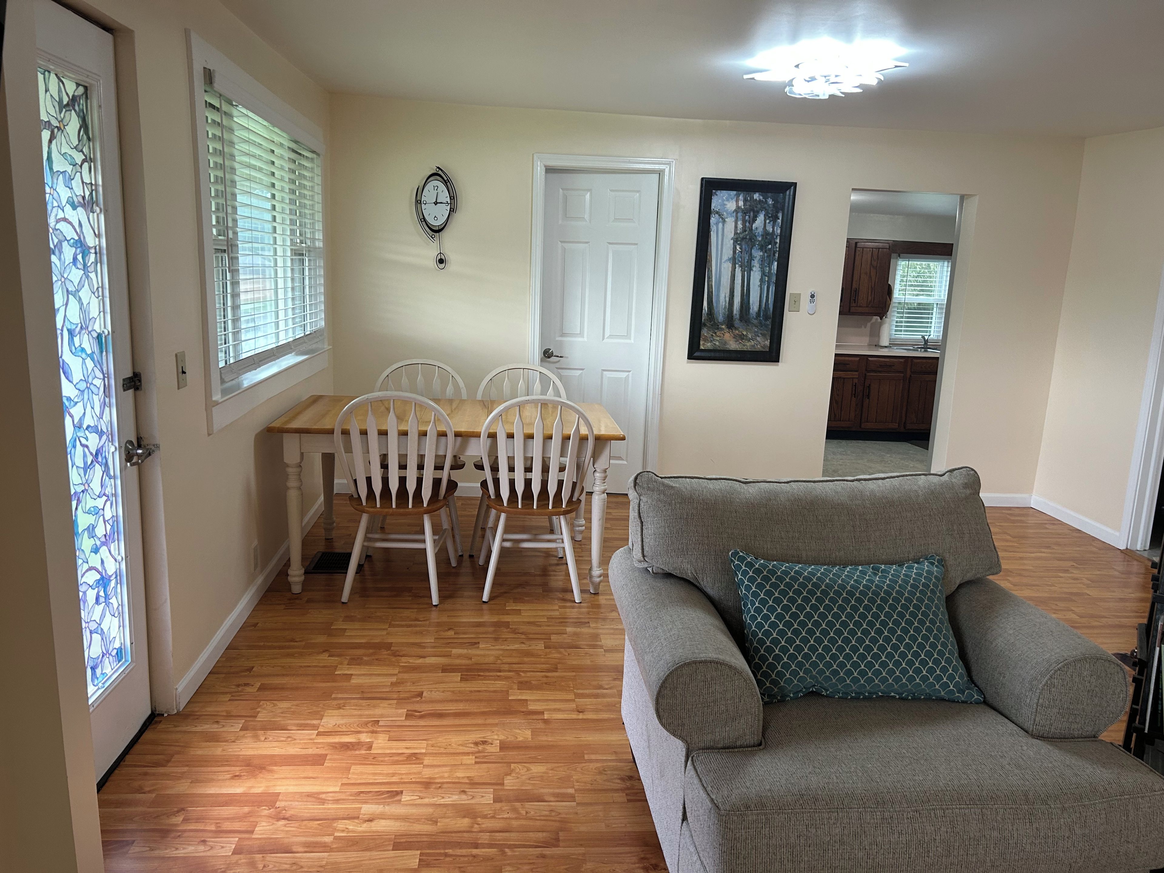 A living room with a grey sofa, a wooden dining table with white chairs, a white door, and a wall clock.