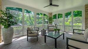 Une terrasse couverte avec un sol en pierre, des fenêtres à cadre blanc et un ventilateur de plafond.