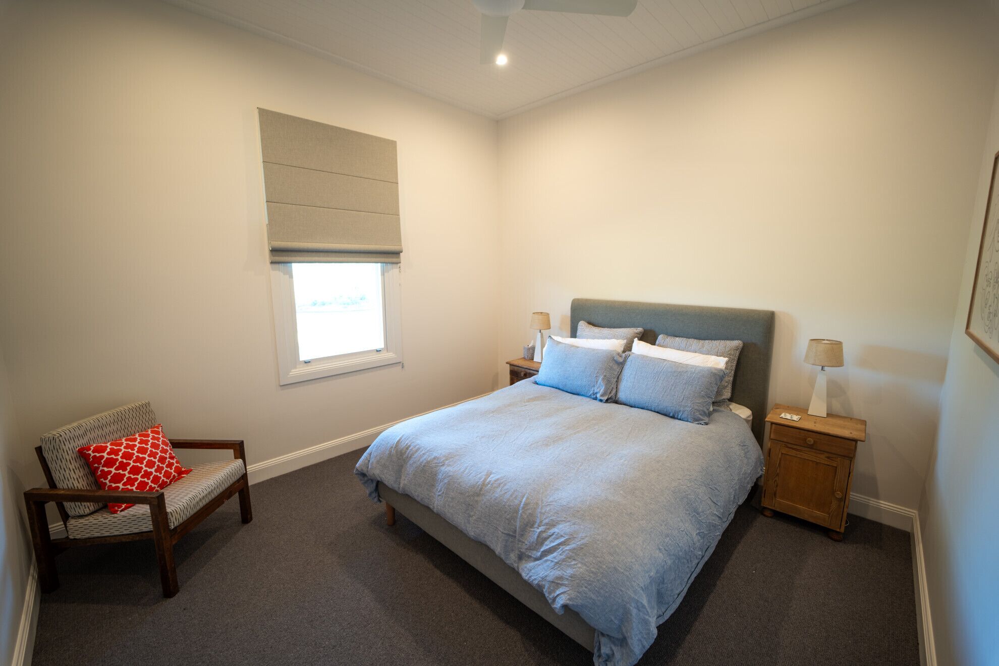 A bedroom with a bed, a chair, a nightstand, a lamp, a window with a blind, and a ceiling fan.