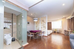 Une chambre d’hôtel moderne dotée d’une paroi en verre séparant la salle de bain, d’un coin repas avec une nappe violette, d’un lit et d’un espace détente avec un ordinateur portable.