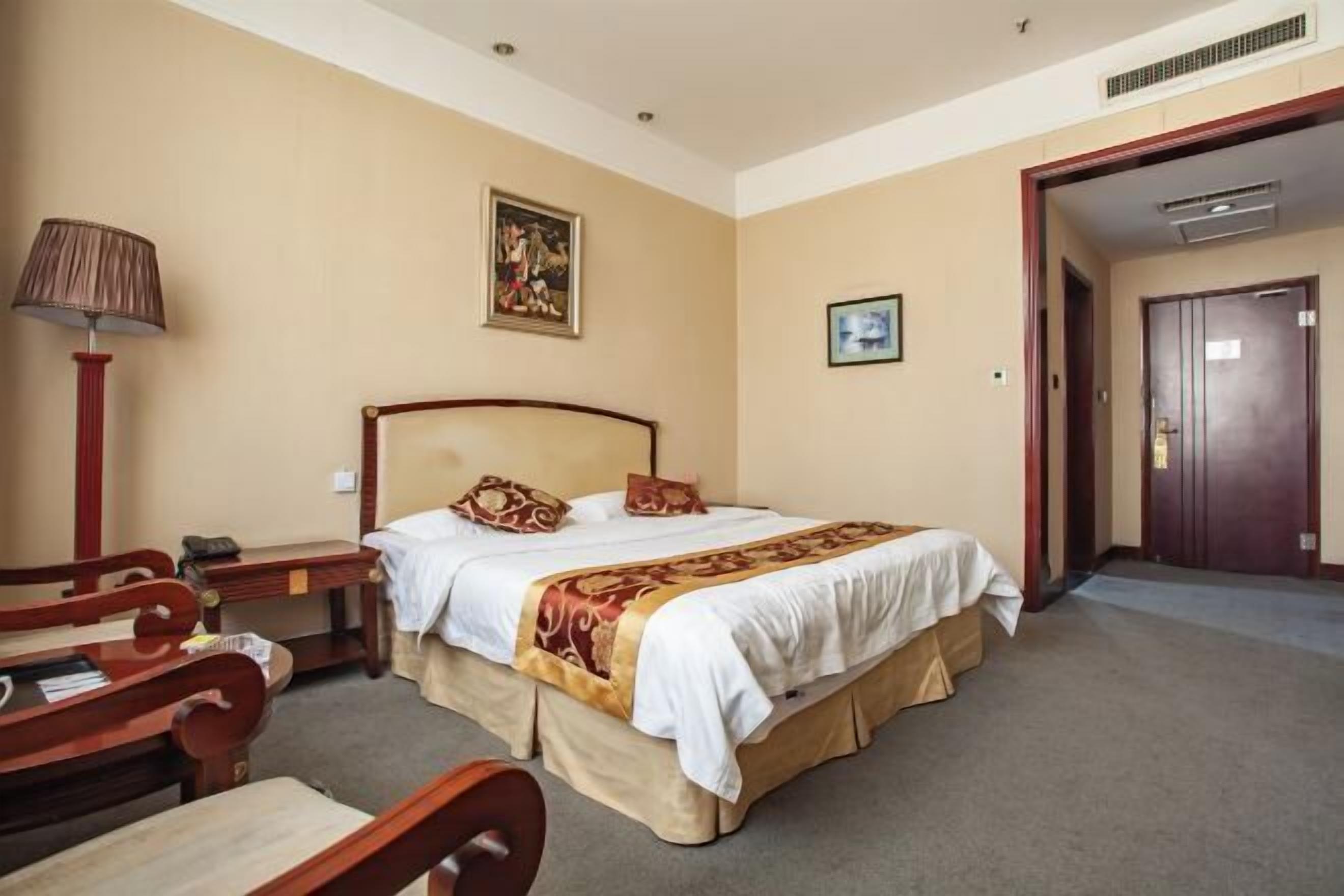 A hotel room with a large bed, a desk with a lamp, and a door leading to another room.