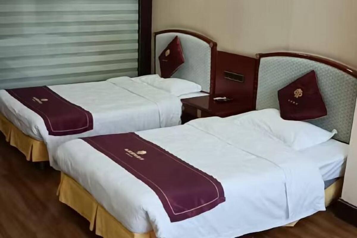 Deux lits simples avec des couvre-lits blancs et bordeaux, des têtes de lit en bois et une table de chevet avec une lampe.