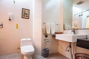 Une salle de bain avec des toilettes, un lavabo et un miroir. On y trouve une poubelle et un porte-serviettes avec des serviettes.