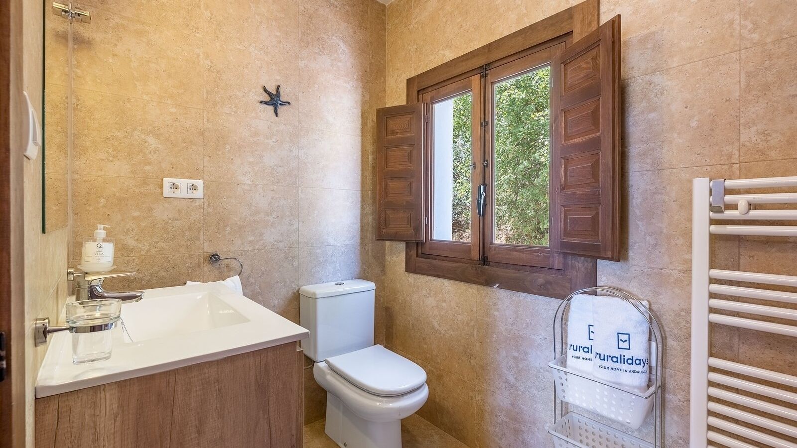Un baño con lavamanos blanco, inodoro, decoración con una estrella de mar, ventana con contraventanas de madera y toallero con un cartel.