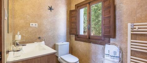 Un baño con lavamanos blanco, inodoro, decoración con una estrella de mar, ventana con contraventanas de madera y toallero con un cartel.