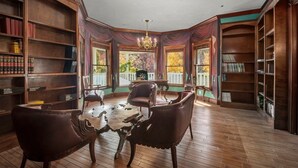 A library with wooden bookshelves, leather chairs, and a large window with a view of trees. - Luxury Mountain Retreat: Skiing, SPA, Arcade & Bar (Wrightwood)