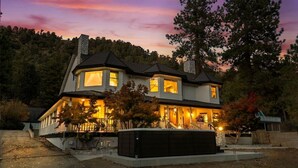 A two-story house with a stone chimney, a porch with railings, and a well-lit front entrance. - Luxury Mountain Retreat: Skiing, SPA, Arcade & Bar (Wrightwood)