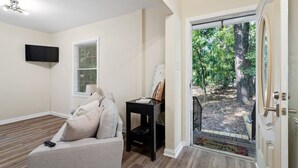 A room with a sofa, a small table with a painting, a TV mounted on the wall, and a door leading to an outdoor area with trees. - Walk Downtown - Private Retreat w King Bed (Fuquay-Varina)