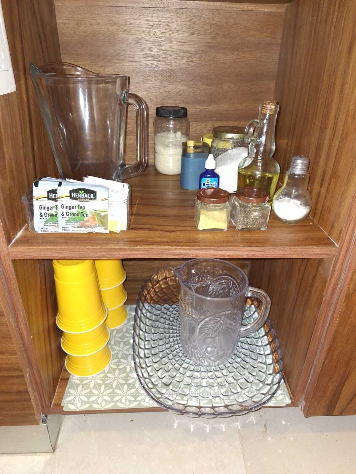 Une étagère de cuisine contenant divers objets, notamment un pichet, des bocaux, une bouteille de Bactérine et une boîte de thé au gingembre.