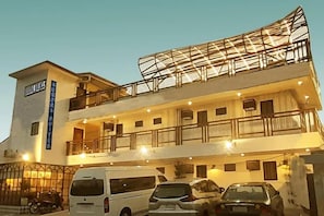 Een meerlaags hotel met een balkon op de bovenste verdieping, een bord met de vermelding dat het een hotel is, en meerdere geparkeerde auto's ervoor.
