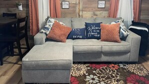 Un sofá seccional gris con dos almohadones, un tapete estampado con motivos florales y una pared de madera con letreros enmarcados.