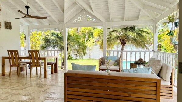 A covered outdoor seating area with wooden furniture, a ceiling fan, and a view of a pool and greenery.