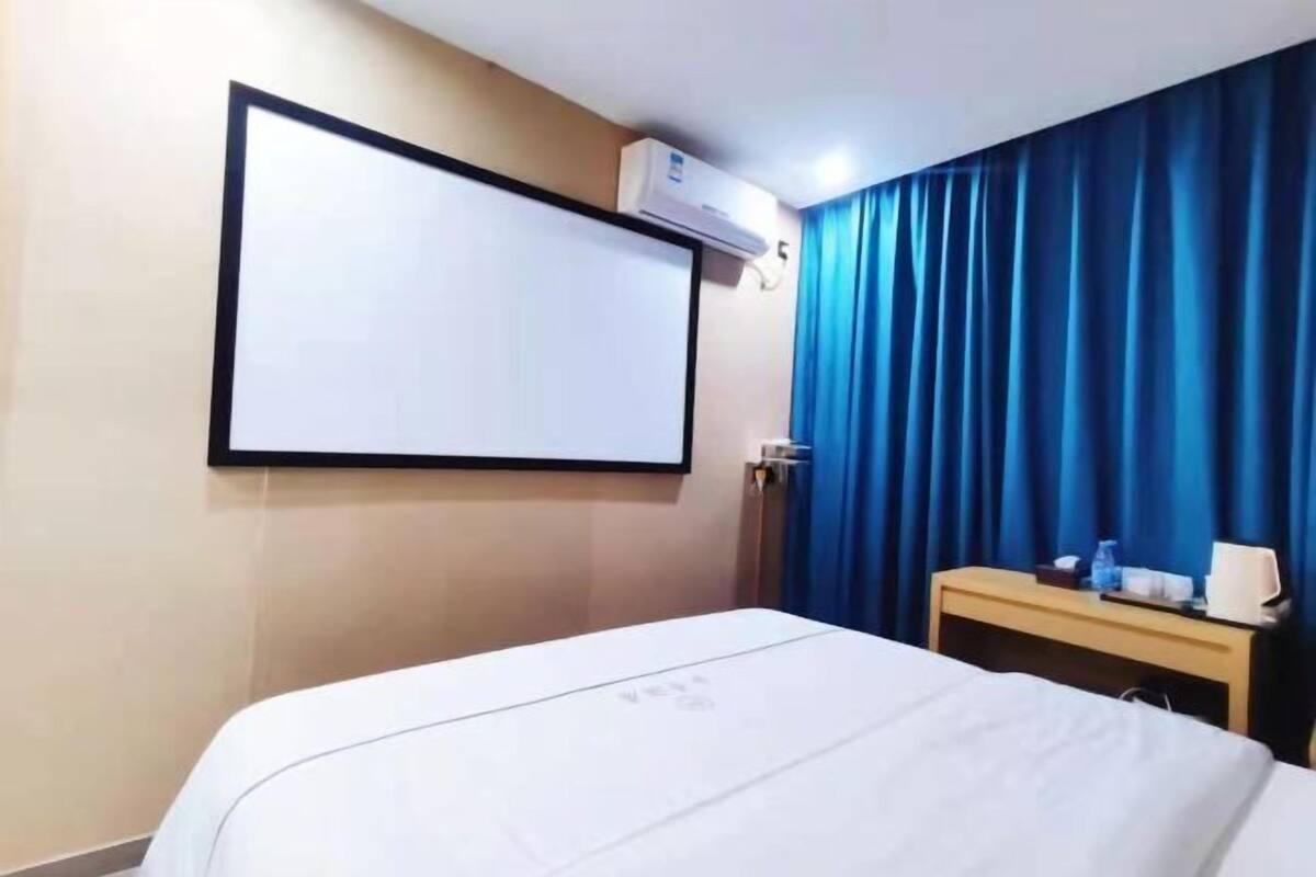 Habitación de hotel con cama, escritorio, aire acondicionado de pared y una ventana grande con cortinas azules.